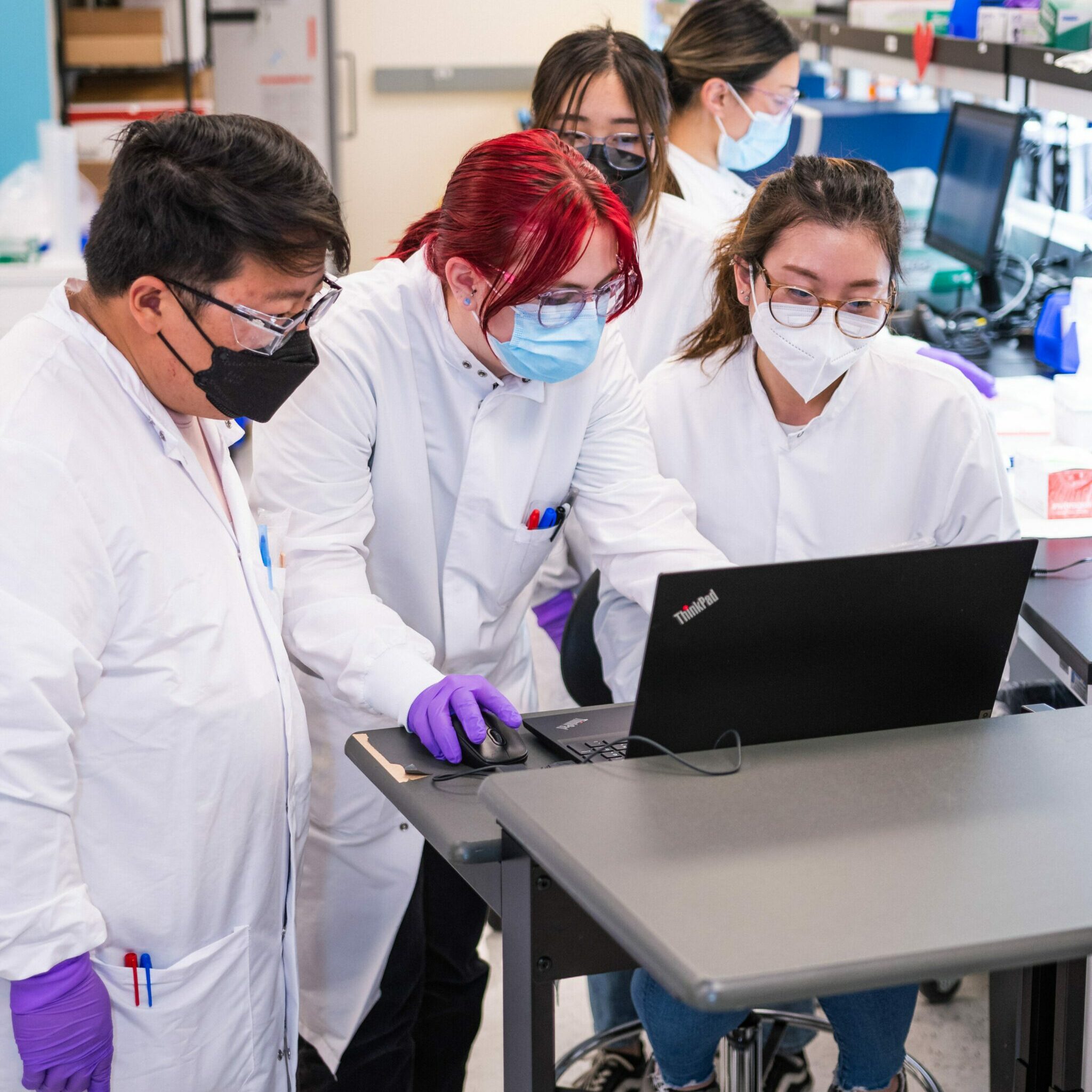 Three employees - a man and two women, one with bright scarlet hair - standing at a computer and working together in a Billion To One lab